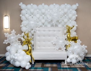 A white Christmas wall display behind a white chair with surrounding white balloon bunches with gold reindeer and white snowflakes.