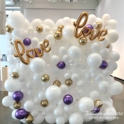 A white balloon wall display with small purple and gold balloons scattered throughout. With two word balloons that say "love".