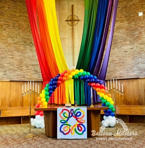 Pride rainbow balloon display with draped rainbow fabrics behind the altar of a church.