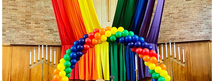 Pride rainbow balloon display with draped rainbow fabrics behind the altar of a church.