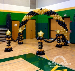 A black and gold balloon arch over a walkway that is strung to a black and gold column on either side.