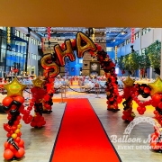 A red, black and gold balloon arch above a red carpet with text "SHA" at the top. There are black and red balloon guardrails along sides of red carpet.