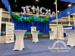 The text "JENCAP" strung in arch above venue surrounded by an assortment of green and cyan balloons.