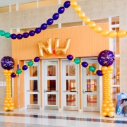 the text "NW" hung above doorway with two gold balloon columns, with a purple Masquerade balloon at the top of each, to the sides of the doorway. there is also green, gold, and purple balloon garlands hung from tall ceilings.