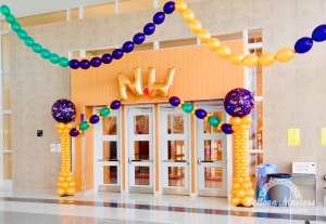 the text "NW" hung above doorway with two gold balloon columns, with a purple Masquerade balloon at the top of each, to the sides of the doorway. there is also green, gold, and purple balloon garlands hung from tall ceilings.