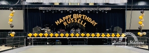 the words "happy birthday russell" strung at back of a stage with the edge of stage lined by black and yellow balloons with two yellow black and marbled balloon bouquets on each side.