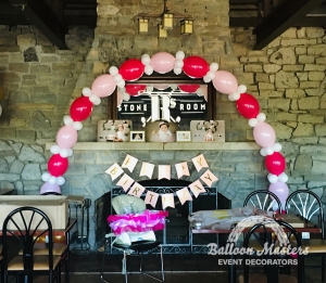 A pink, red and white balloon arch over stone fireplace.