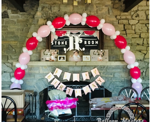 A pink, red and white balloon arch over stone fireplace.