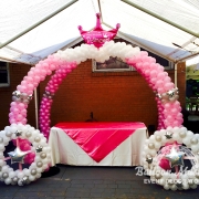 Two hombre pink to white balloon arches that create a cross formation with a pink princess crown balloon at top. There are also two silver and white balloon wreaths in the front.