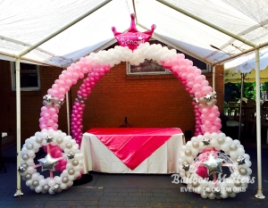 Two hombre pink to white balloon arches that create a cross formation with a pink princess crown balloon at top. There are also two silver and white balloon wreaths in the front.