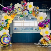 A 60s themed rainbow marbled balloon arch with big white daisies, tie dye peace signs, and barbie balloons.
