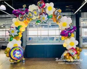 A 60s themed rainbow marbled balloon arch with big white daisies, tie dye peace signs, and barbie balloons.