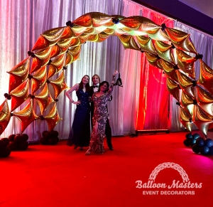 A gold and black, wide balloon arch created from staggered star balloons over a red carpet.