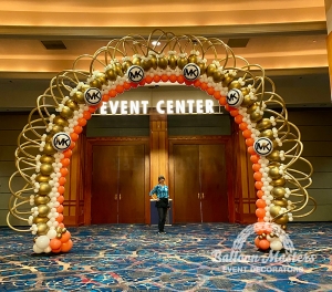 A large, gold, white and orange balloon arch with Michael Kors logos evenly spaced on it.