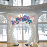 a white balloon arch shaped to look like a unicorn with a gold horn and pink, purple, and blue hair.