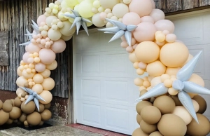 Brown, orange, peach, and yellow balloon arch with silver stars attached infront of garage.