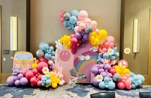 Purple, pink, blue, and yellow balloon display with balloon flowers, disco balls and 60s cut outs scattered in balloons.